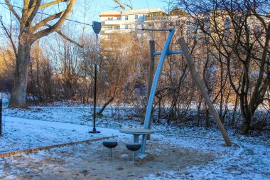 Bir salıncak seti ve dairesel oturma alanı kışın ağaçlar ve güneş ışığıyla çevrili karlı bir parkta duruyor..