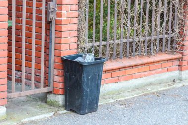Banliyö kaldırımında dövülmüş demir çitli tuğladan bir evin önüne konmuş siyah plastik çöp kutusu.