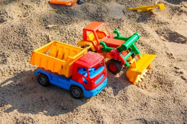 Ön yükleyicili iki renkli plastik oyuncak kamyonu çocukların kum havuzu bölgesinde birlikte çalışıyor.