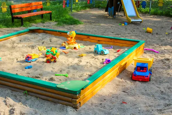 Kare tahta kum havuzu oyuncaklarla dolu ve bahçedeki yeşil çimlerle çevrili.