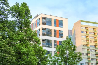 Çevresi yemyeşil ağaçlarla çevrili, ayırt edici turuncu aksanlı modern apartman binası.