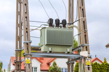 Beton elektrik direğine monte edilmiş elektrik trafosu. Arkaplanı temiz gökyüzünün altında görünen konut binaları var.