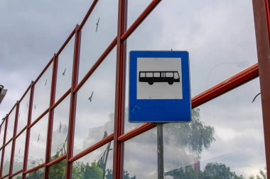 Şehir merkezinde bulutlu gökyüzüne karşı kırmızı metal parmaklıklara monte edilmiş mavi dikdörtgen otobüs durağı tabelası.