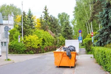 Yeşil ağaçlar ve çitlerle kaplı sessiz bir banliyö caddesinde parlak turuncu inşaat atığı konteynırı