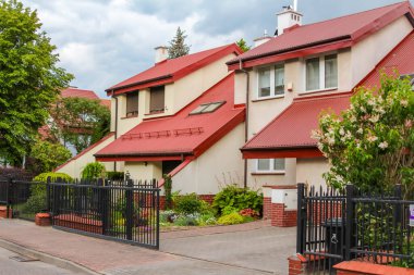 Güneşli bir günde, Kırmızı Döşemeli Çatı, Siyah Metal Çit ve Peyzaj Bahçeli Modern Banliyö Evi