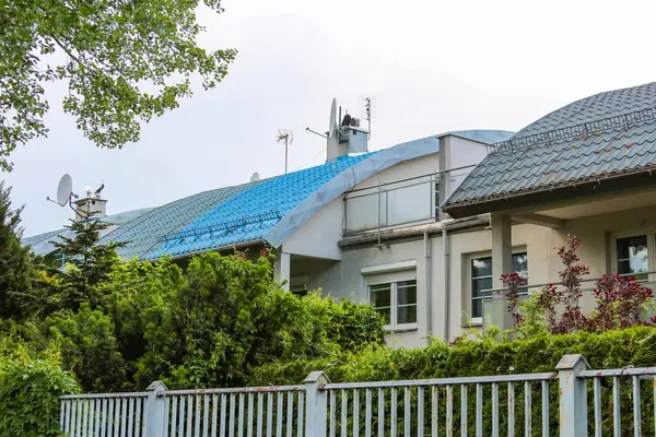 Beyaz çitli modern banliyö evi ve bulutlu gökyüzünün altında mavi sanayi binasının yakınındaki bahçe manzarası.