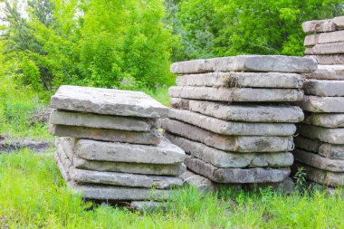 Parlak gökyüzünün altında yeşil ağaçlarla çevrili çimenli bir alanda büyük beton bloklar yığılmış.,