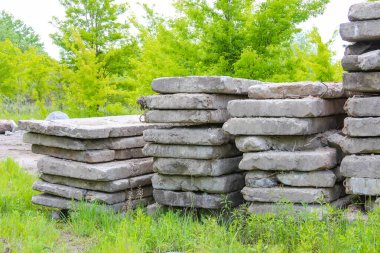 Büyük, yıpranmış beton bloklar çimenli bir tarlada düzgünce istiflenmiş, arka planda yeşil ağaçlar, inşaat malzemelerini gösteriyor.,