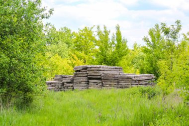 Parlak bir gökyüzünün altında, yemyeşil çimenler üzerinde istiflenmiş eski ahşap kirişlerin bulunduğu bir açık hava sahnesi.,