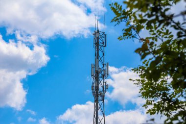 Telekomünikasyon kulesi, arka planda mavi gökyüzü ve beyaz bulutlar ile yeşil ağaç dalları üzerinden görülüyor.
