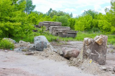 Büyük beton bloklar ve kayalar kumlu, toprak ve seyrek yeşil bitki örtüsü üzerine dağılmış. Bu da terk edilmiş bir inşaat sahası ya da doğal bir manzarayı işaret ediyor.,