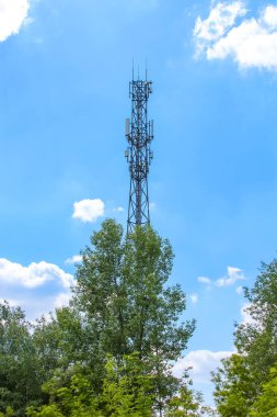 Uzun, ince bir hücresel iletişim kulesi, açık mavi gökyüzüne karşı duruyor. Dağılmış bulutlar, yeşil ağaçlarla çevrili, modern teknolojiyi sembolize ediyor.,