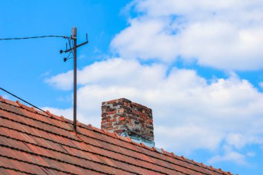Geleneksel turuncu kiremit çatılı, tuğla bacalı ve antenli, açık mavi gökyüzüne karşı kurulmuş.,