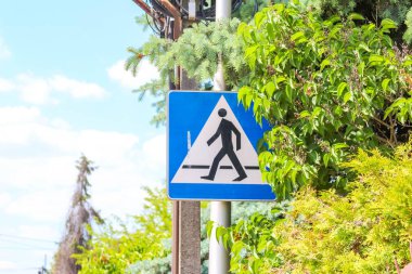 İkamet alanındaki yemyeşil yeşilliklere karşı metal direğe monte edilmiş mavi üçgen yaya geçidi uyarı levhası.