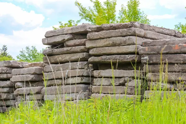 Büyük beton levhalar çimenli bir arazide düzenli bir şekilde istiflenir. Arkaplanda yeşil ağaçlar vardır. Bu da inşaat ve depolama alanlarını gösterir.,