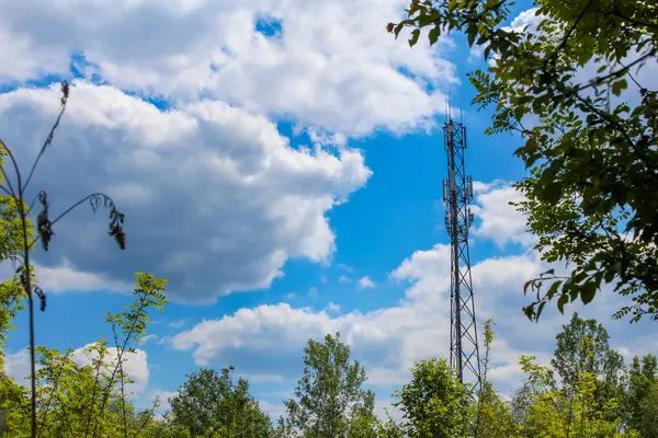 Haberleşme kulesi, yeşil orman tepe örtüsünün üstünde mavi gökyüzüne karşı dağınık beyaz bulutlarla yükseliyor.