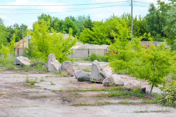 Büyük yıpranmış beton bloklar ve kırık taş levhalar parlak bir gökyüzünün altında seyrek ağaçlarla kaplı çimenli bir alana saçılmış. Bu da terk edilmiş bir inşaat sahası veya doğal bir manzarada kalıntılar olduğunu gösteriyor.,