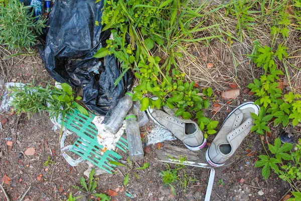 Torbalar ve bilinmeyen parçalar da dahil olmak üzere çeşitli beyaz ve siyah plastik çöp parçaları bir toprak ve çimen arazisine saçılmış, çevresel kirlilik ve dikkatsiz atık imhası vurgulanmıştır.,