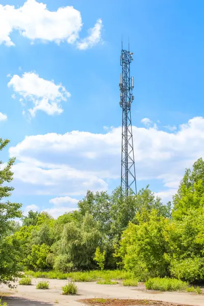 Kırsal alanda bulutlu mavi gökyüzüne karşı yeşil ağaçların arasında dimdik duran iletişim kulesi