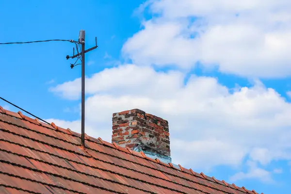 Geleneksel turuncu kiremit çatılı, tuğla bacalı ve antenli, açık mavi gökyüzüne karşı kurulmuş.,