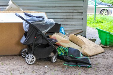 Evin duvarına park etmiş, yanında bahçe aletleri ve yeşil bir kova olan siyah bebek arabası.