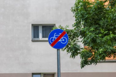 Beyaz köşegen çizgili mavi dairesel trafik levhası gri bina duvarına yaslanmış yeşillik ile