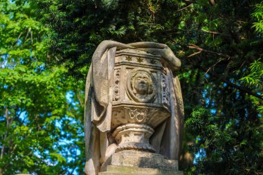 Yıpranmış taş heykel mezarlık bahçesinde yoğun yeşil bitki örtüsüyle çevrili.