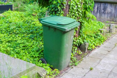 Konut alanındaki bahçe çitinin yakınındaki beton kaldırımda duran yeşil plastik atık kutusu