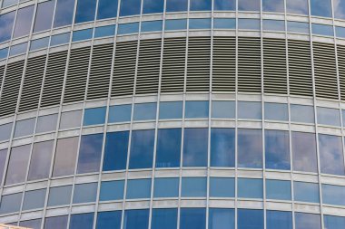 Modern bir gökdelenin cam ve metal cephesinin yakın görüntüsü, yinelenen pencere ve yansımaları gösteriyor.