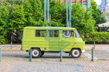 Yeşil ağaçların yanındaki asfalt bir yüzeye park etmiş, üzerinde sarı yedek lastiği olan eski, açık yeşil bir minivan..