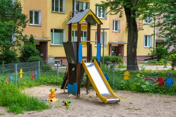 Parkta yeşil ağaçlarla çevrili sarı kaydırak ve tırmanma yapısına sahip oyun alanı ekipmanları