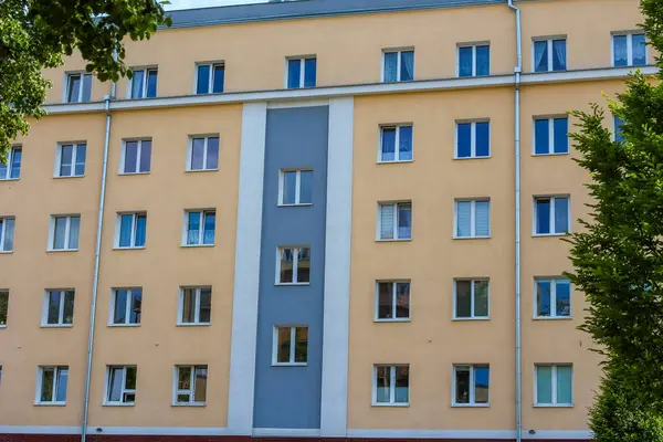 Yeşil ağaçlarla çevrili mavi pencereli ve birden fazla katlı büyük bej apartman dairesi.