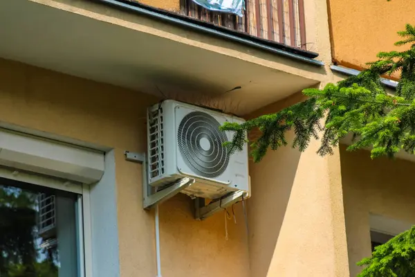 Pencerenin yanındaki sarı duvara monte edilmiş havalandırma ünitesi ve yeşil yapraklı bina köşesi.