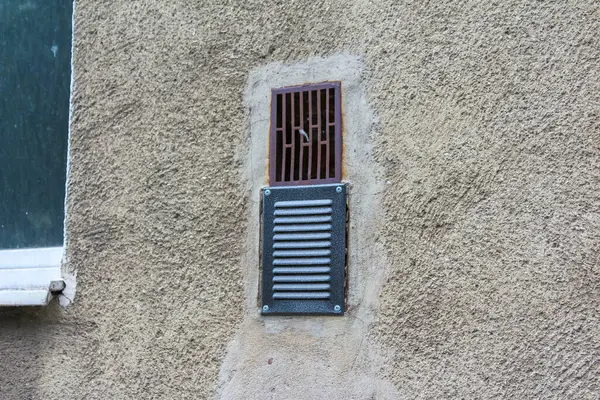 Metal ızgaralı, küçük, parmaklıklı bir pencere. Kalıplı, açık kahverengi bir bina duvarına yerleştirilmiş. Güvenlik veya bodrum katı olabilir..