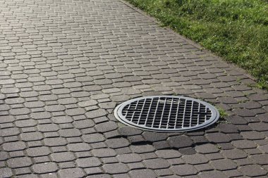 Çakıl taşı kaldırımına gömülü geometrik ızgara desenli yuvarlak rögar kapağı kentsel altyapı detaylarını oluşturuyor