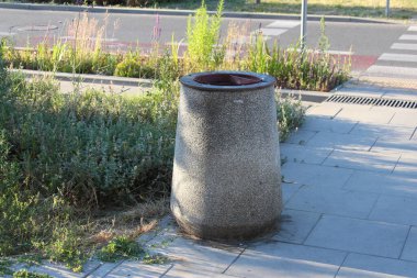 Yeşil bitkilerle dolu şehir sokağının yanındaki kaldırıma yerleştirilmiş büyük beton silindirik atık kutusu