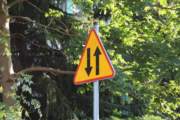 Sarı üçgen uyarı levhası, yeşil yapraklarla çevrili metal direğe takılı ünlem işareti.