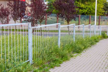 Asfalt kaldırım boyunca uzanan uzun beyaz metal çit. Ağaçlar ve yerleşim alanı arka planında.