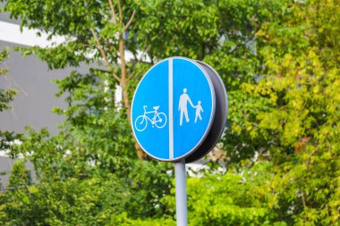 Yeşil yapraklar arasında direğe monte edilmiş yaya ve bisikletçi sembollü mavi dairesel trafik tabelası