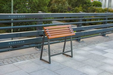 Metal korkulukların yanına kurulmuş boş ahşap park bankı gündüz boyunca huzurlu kentsel yeşil alana bakıyor