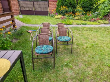 İkamet binası ve peyzajın yanında yeşil çimlere dizilmiş renkli yastıklı iki metal bahçe sandalyesi.