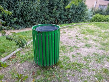 Doğal bitki örtüsü ve kentsel gelişim geçmişine sahip yerleşim alanındaki kumlu zemine yerleştirilmiş parlak yeşil silindirik atık konteynırı