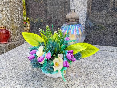 Granit mezarlık yüzeyindeki anıtın yanına yerleştirilmiş mor ve yeşil suni çiçek düzenlemesi düşünceli bir haraç kompozisyonu sergiliyor.