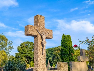 Yeşil yapraklı, berrak mavi gökyüzüne karşı duran taş haçı heykel huzur dolu mezarlık manzarası yaratıyor.