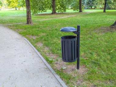 Yeşil şehir parkında kavisli yürüyüş yolunun yanında duran kamu atığı, şehrin temel altyapı hizmetlerini sunuyor.