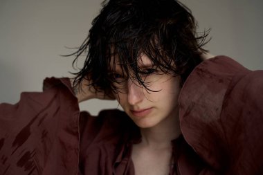 Burgonya kapüşonlu, ıslak saçlı genç, duygusal portresiyle yüzüne dokunuyor.