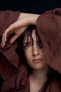 Terracotta renkli kapüşonlu esmer kadın portresi. Yüzüne yakın el hareketleriyle.