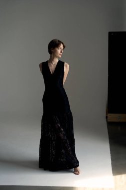 Siyah gece elbisesi içinde zarif bir kadın stüdyo ortamında modern mimari arka planda poz veriyor.