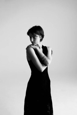 Kolsuz, siyah bir gece giymiş zarif bir kadın. Stüdyoda yüz yüze düşünceli bir şekilde poz veriyor.