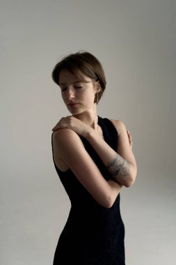 Siyah kolsuz elbiseli, kollarını kavuşturup doğrudan kameraya bakan kendinden emin genç bir kadın.